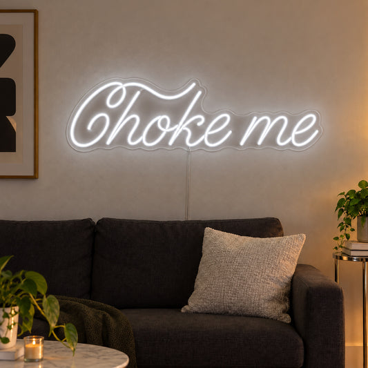 Choke Me Neon Sign in white, featuring Choke Me script lettering above a dark sofa.
