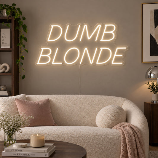 Warm White version of the Dumb Blonde Neon Sign, shown in slanted lettering above a cream sofa.