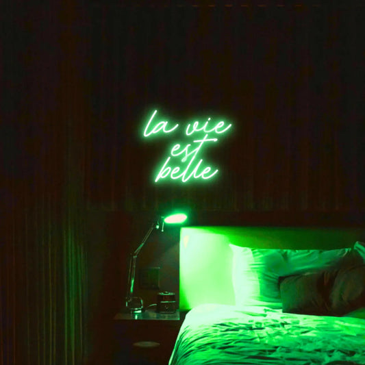 La Vie est Belle LED Neon Light Sign in green, featuring script lettering illuminated above a bed.