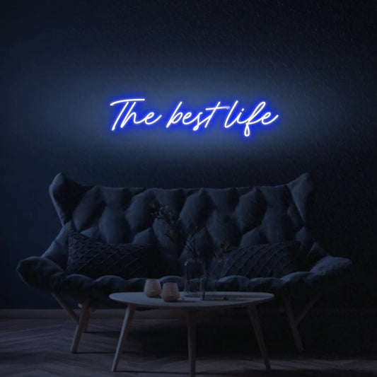 The Best Life LED Neon Light Sign in blue, featuring script lettering illuminated above a dark tufted sofa.