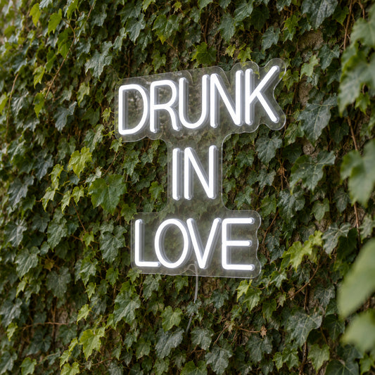 Drunk in Love LED Neon Light Sign in white, featuring three lines of block lettering against a green foliage wall.