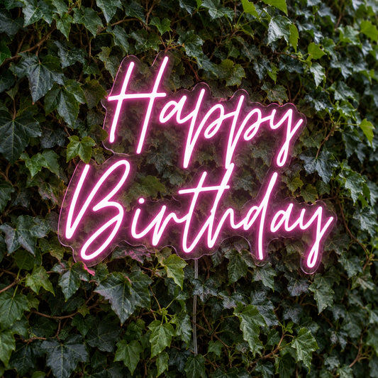 Happy Birthday LED Neon Light Sign in pink, featuring two lines of script lettering against a green foliage wall.