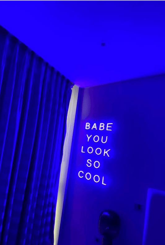 Babe You Look So Cool 2 LED Neon Light Sign in purple, featuring five stacked words on a bedroom wall beside curtains.