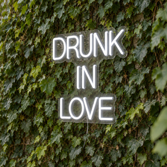 Drunk in Love LED Neon Light Sign in white, featuring three lines of block lettering against a green foliage wall.