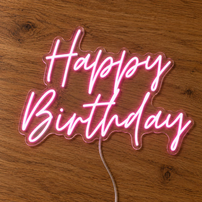 Happy Birthday LED Neon Light Sign in pink, featuring two lines of script lettering on a wooden surface.