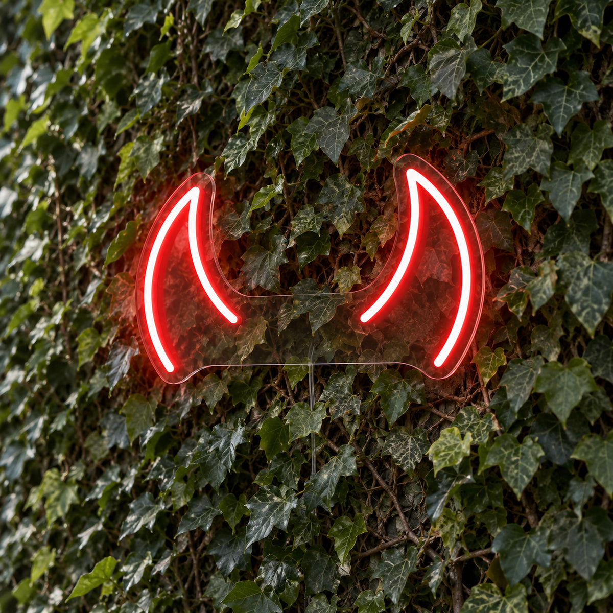 Horns LED Neon Light Sign in red, featuring pair of devil-horn outlines against a green foliage wall.
