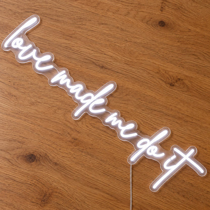 Love Made Me Do It LED Neon Light Sign in white, featuring script lettering on a wooden surface, viewed at an angle.