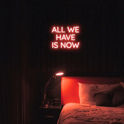 All We Have Is Now LED Neon Light Sign in red, featuring three lines of block lettering illuminated above a bed.