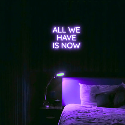 All We Have Is Now LED Neon Light Sign in purple, featuring three lines of block lettering illuminated above a bed.