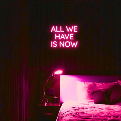 All We Have Is Now LED Neon Light Sign in pink, featuring three lines of block lettering illuminated above a bed.