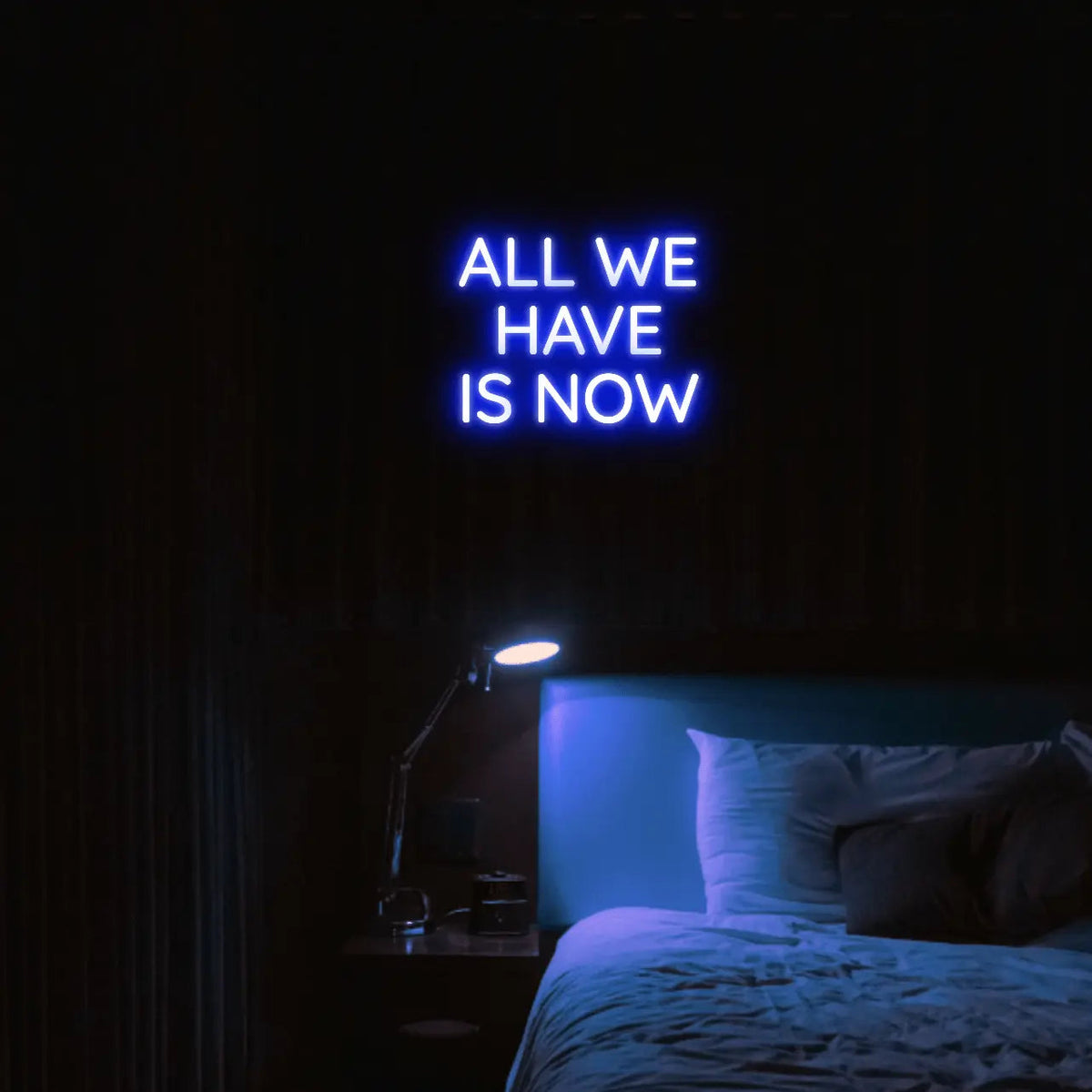 All We Have Is Now LED Neon Light Sign in blue, featuring three lines of block lettering illuminated above a bed.