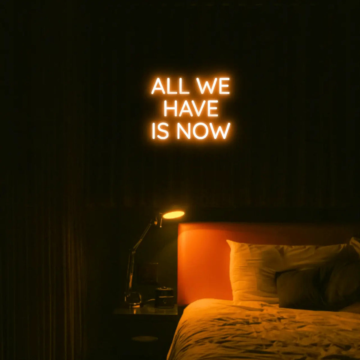 All We Have Is Now LED Neon Light Sign in orange, featuring three lines of block lettering illuminated above a bed.
