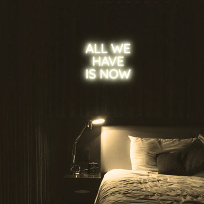 All We Have Is Now LED Neon Light Sign in warm white, featuring three lines of block lettering illuminated above a bed.