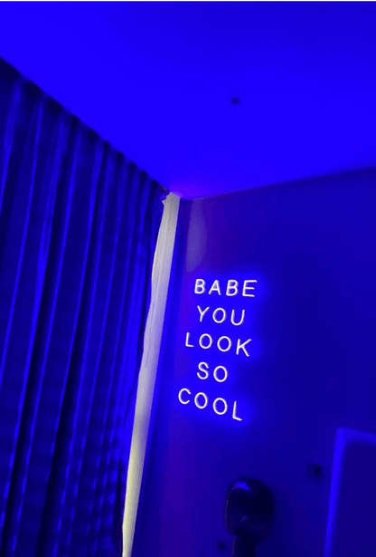 Babe You Look So Cool 2 LED Neon Light Sign in purple, featuring five stacked words on a bedroom wall beside curtains.