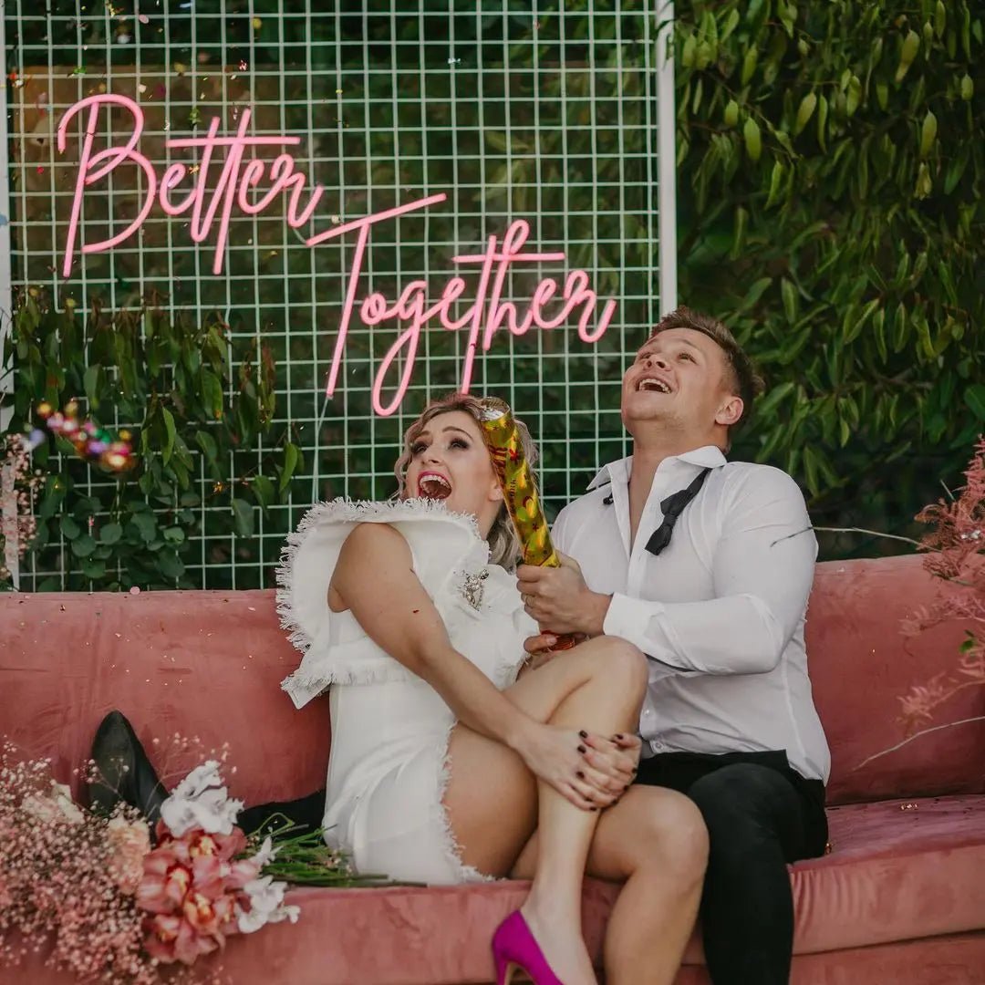 Better Together LED Neon Light Sign in pink, featuring script lettering on a wire-grid wedding backdrop, shown above a couple on a sofa.