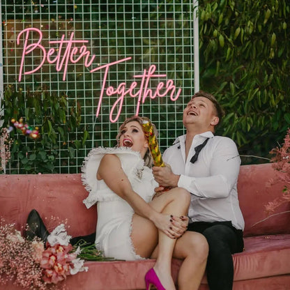 Better Together LED Neon Light Sign in pink, featuring script lettering on a wire-grid wedding backdrop, shown above a couple on a sofa.