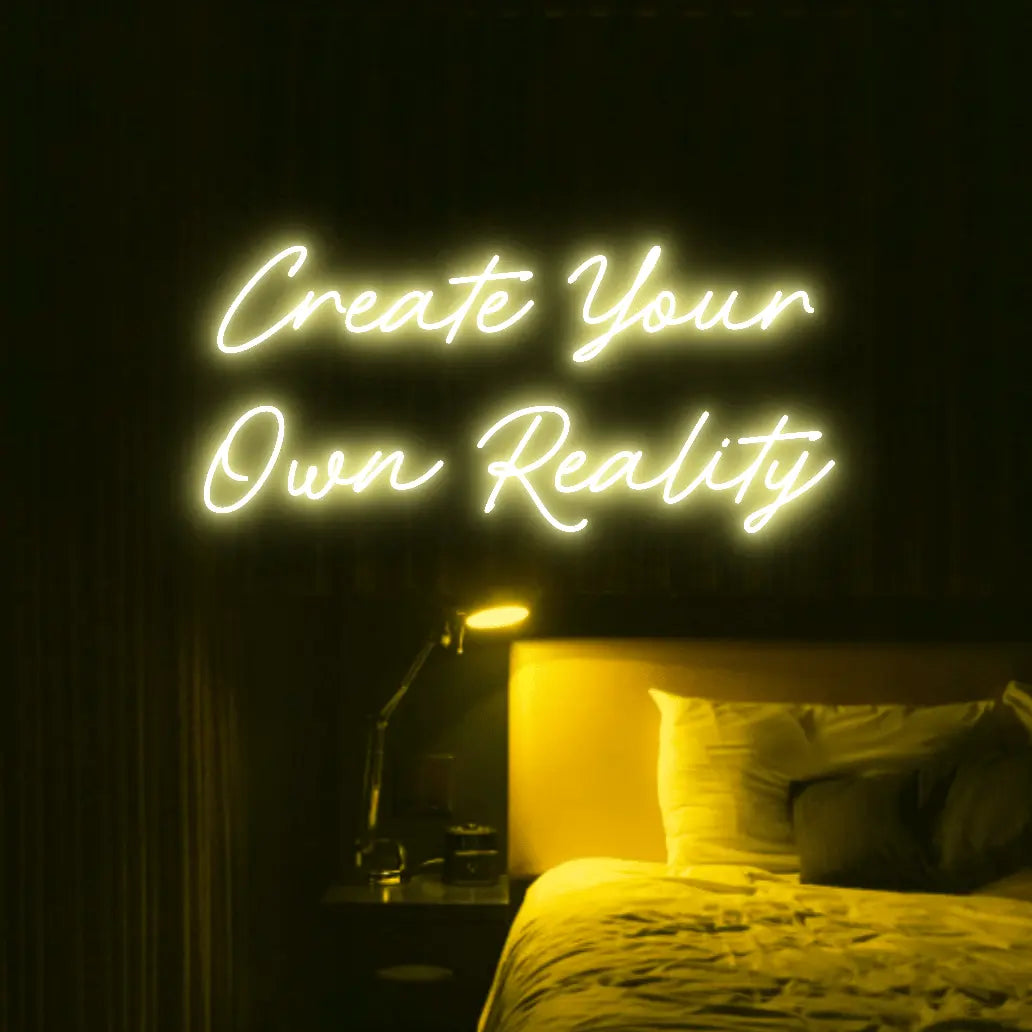 Create Your Own Reality LED Neon Light Sign in yellow, featuring two lines of script lettering illuminated above a bed.