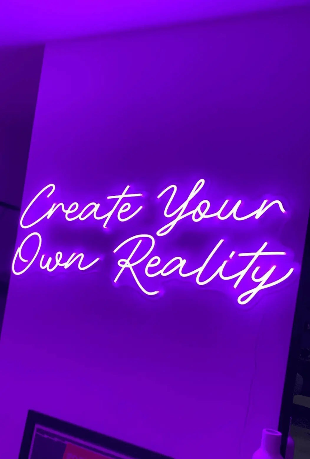 Create Your Own Reality LED Neon Light Sign in purple, featuring two lines of script lettering on a purple-lit wall.