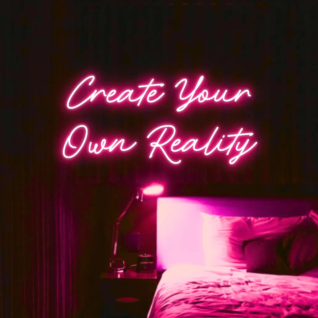Create Your Own Reality LED Neon Light Sign in pink, featuring two lines of script lettering illuminated above a bed.