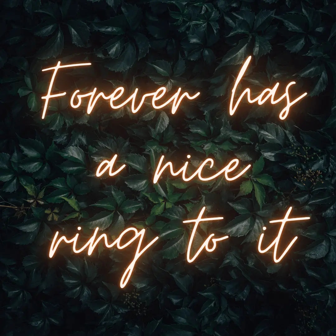 Forever Has a Nice Ring to It LED Neon Light Sign in orange, featuring three lines of script lettering against a dark foliage backdrop.