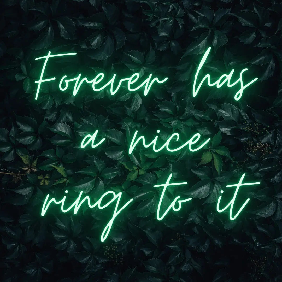 Forever Has a Nice Ring to It LED Neon Light Sign in green, featuring three lines of script lettering against a dark foliage backdrop.