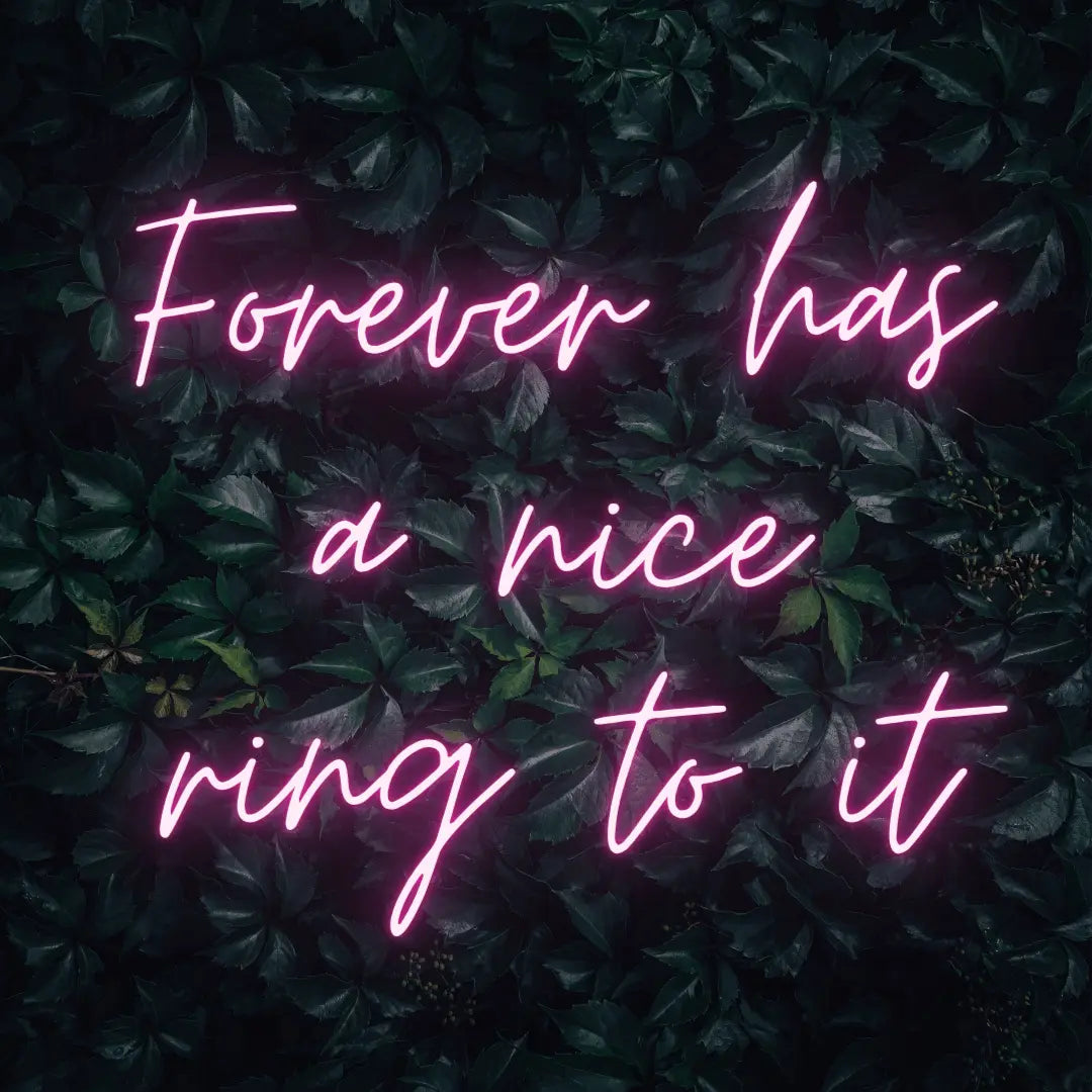 Forever Has a Nice Ring to It LED Neon Light Sign in pink, featuring three lines of script lettering against a dark foliage backdrop.