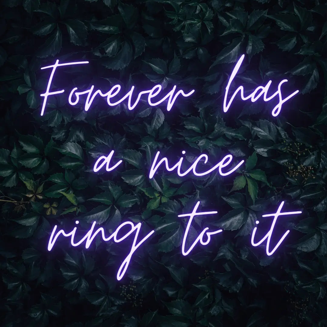 Forever Has a Nice Ring to It LED Neon Light Sign in purple, featuring three lines of script lettering against a dark foliage backdrop.