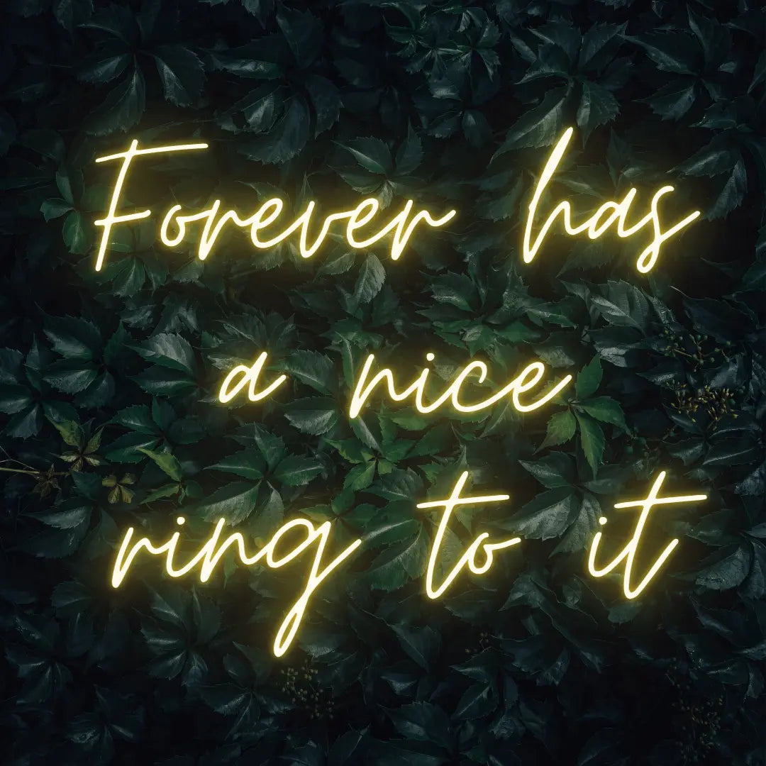 Forever Has a Nice Ring to It LED Neon Light Sign in yellow, featuring three lines of script lettering against a dark foliage backdrop.