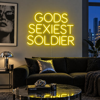 Gods Sexiest Soldier Neon Sign with yellow lighting, displayed in rounded lettering above a dark sofa.