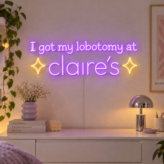 I Got My Lobotomy At Claire's Neon Sign with purple lettering with two yellow sparkle shapes on transparent acrylic, displayed on a living room wall