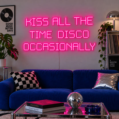Rose Red version of the Kiss All The Time Disco Occasionally Neon Sign, shown above a blue sofa near a disco ball.