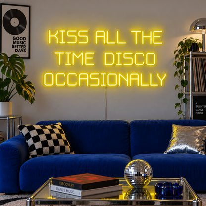 Kiss All The Time Disco Occasionally Neon Sign with yellow lighting, displayed above a blue sofa near a disco ball.
