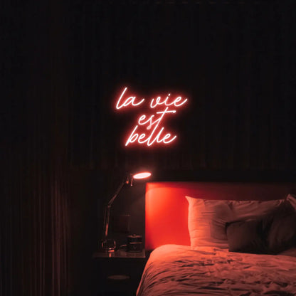 La Vie est Belle LED Neon Light Sign in red, featuring script lettering illuminated above a bed.