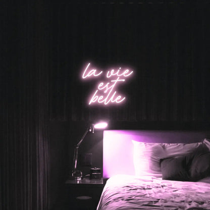 La Vie est Belle LED Neon Light Sign in pale pink, featuring script lettering illuminated above a bed.