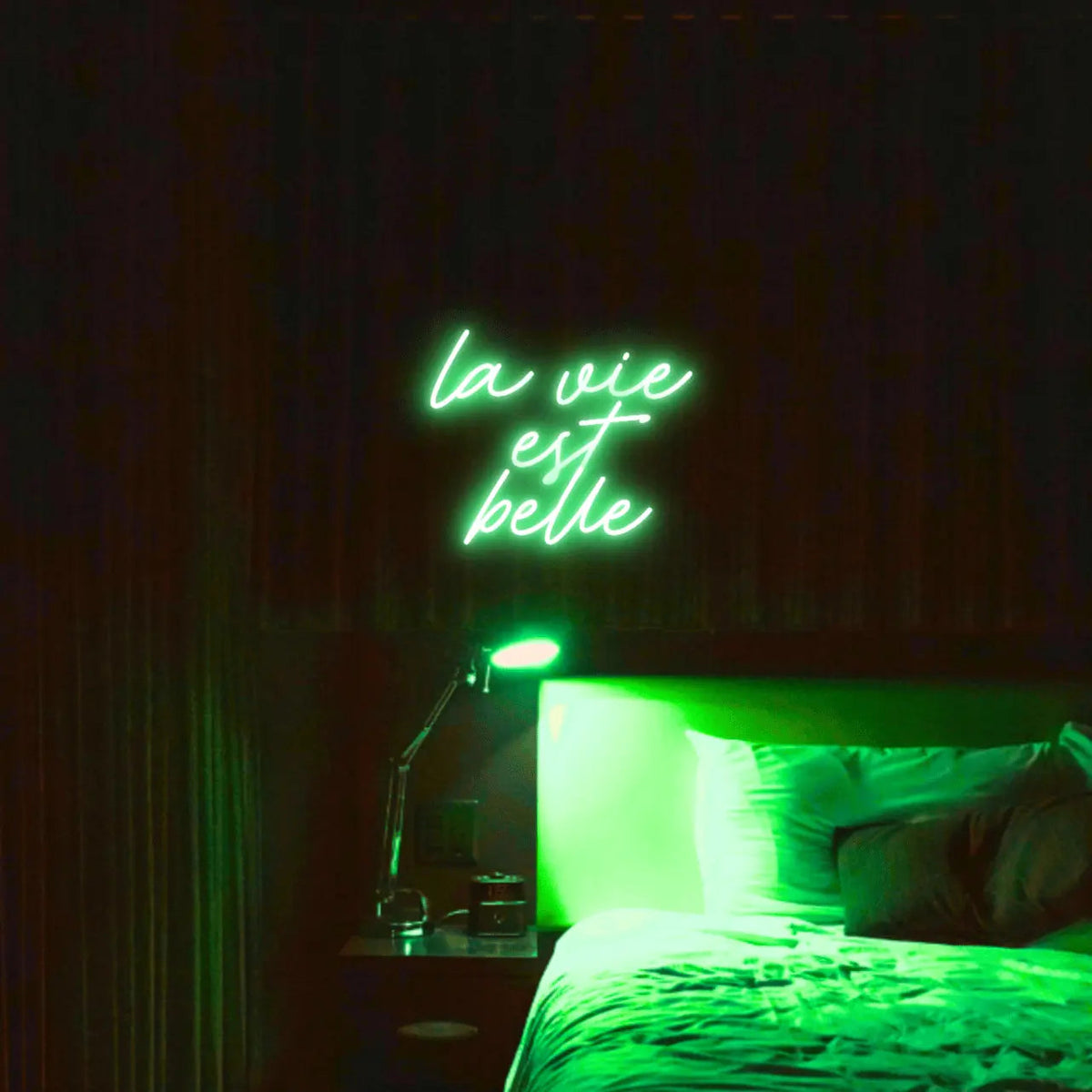 La Vie est Belle LED Neon Light Sign in green, featuring script lettering illuminated above a bed.