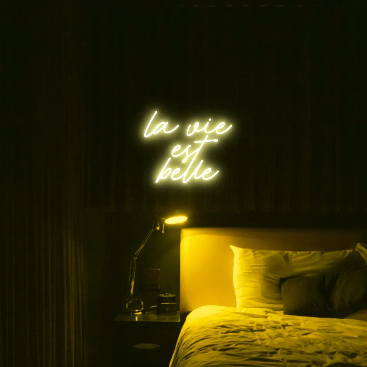 La Vie est Belle LED Neon Light Sign in yellow, featuring script lettering illuminated above a bed.