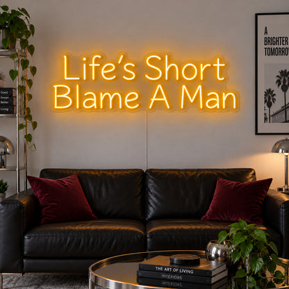 Life's Short Blame A Man Neon Sign glowing gold above a black leather sofa.