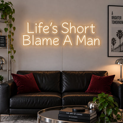 Warm White version of the Life's Short Blame A Man Neon Sign, shown above a black leather sofa.