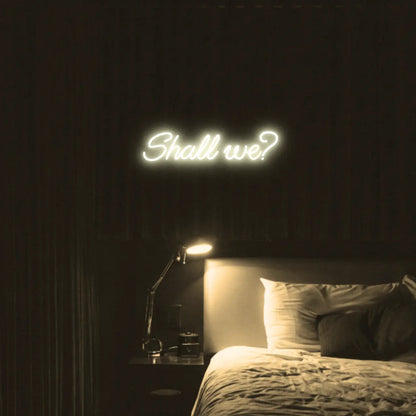 Shall we? LED Neon Light Sign in warm white, featuring script lettering illuminated above a bed.