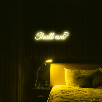 Shall we? LED Neon Light Sign in yellow, featuring script lettering illuminated above a bed.
