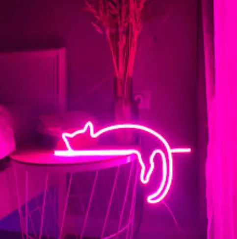 Sleeping Cat LED Neon Sign in pink, featuring sleeping-cat outline illuminated beside a table in a dark room.