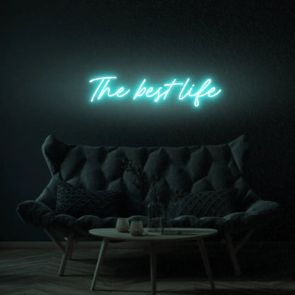 The Best Life LED Neon Light Sign in turquoise, featuring script lettering illuminated above a dark tufted sofa.