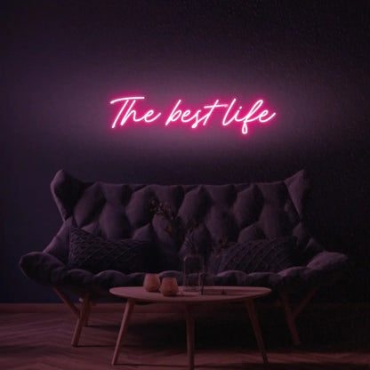 The Best Life LED Neon Light Sign in pink, featuring script lettering illuminated above a dark tufted sofa.