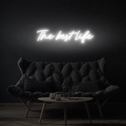 The Best Life LED Neon Light Sign in white, featuring script lettering illuminated above a dark tufted sofa.