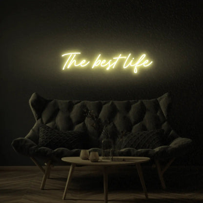 The Best Life LED Neon Light Sign in yellow, featuring script lettering illuminated above a dark tufted sofa.