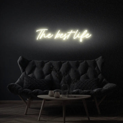 The Best Life LED Neon Light Sign in warm white, featuring script lettering illuminated above a dark tufted sofa.