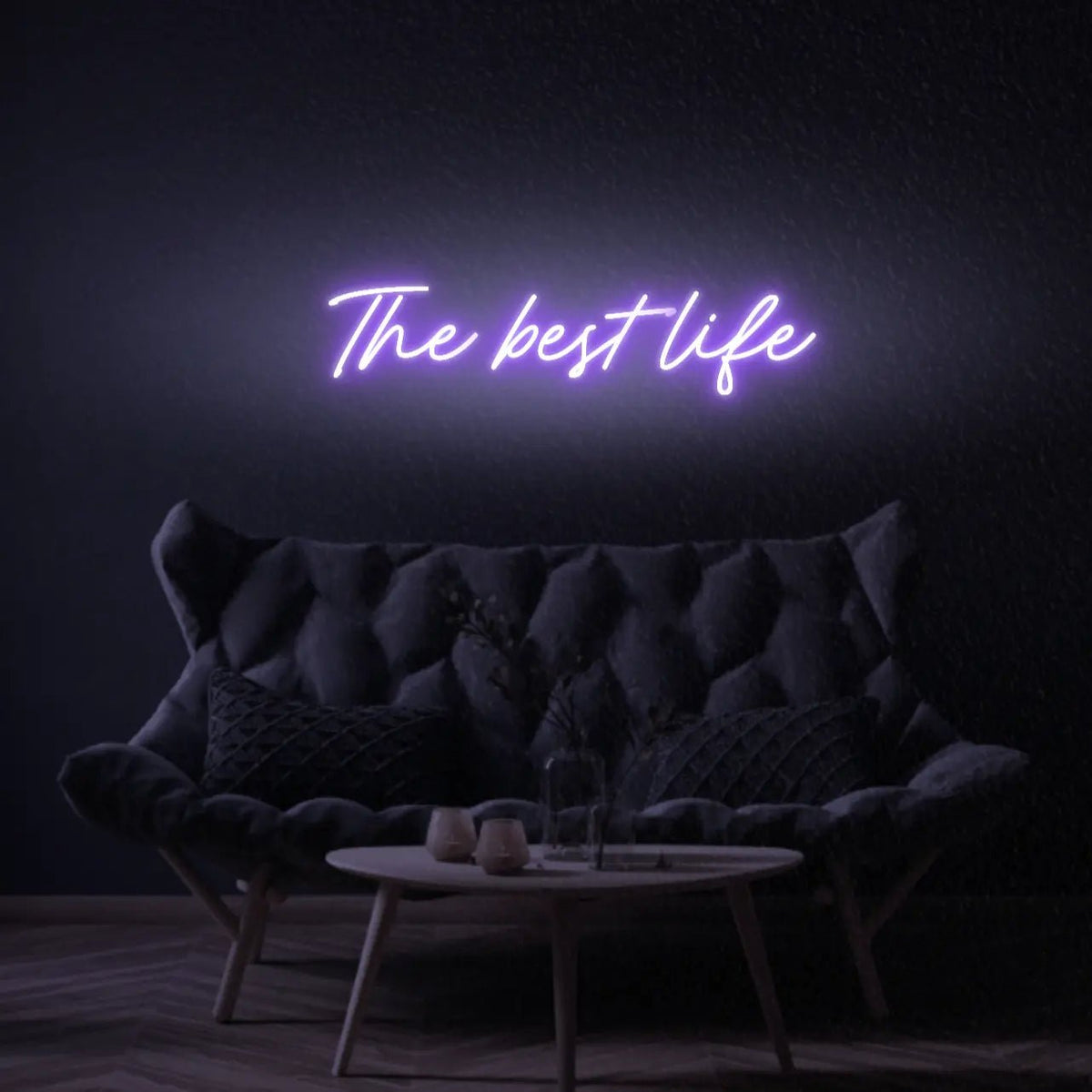 The Best Life LED Neon Light Sign in purple, featuring script lettering illuminated above a dark tufted sofa.