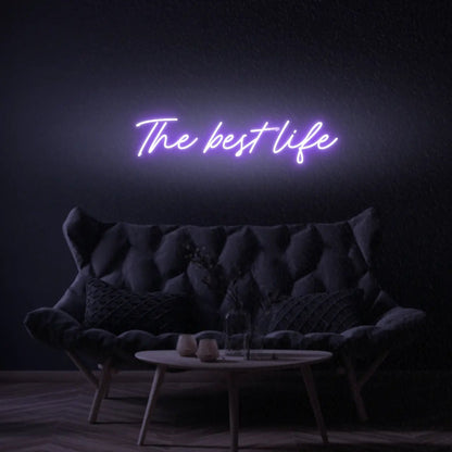 The Best Life LED Neon Light Sign in purple, featuring script lettering illuminated above a dark tufted sofa.