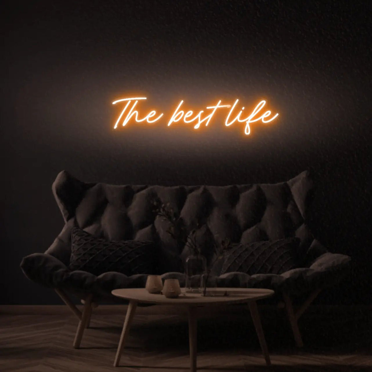 The Best Life LED Neon Light Sign in orange, featuring script lettering illuminated above a dark tufted sofa.