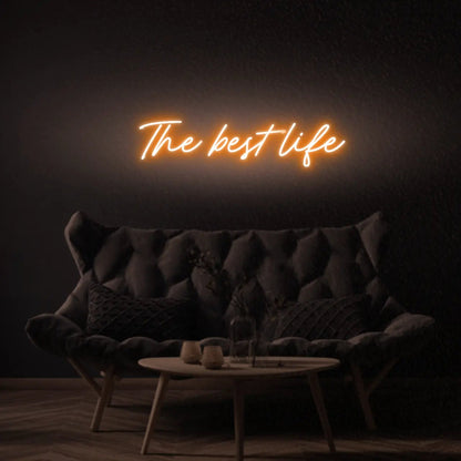 The Best Life LED Neon Light Sign in orange, featuring script lettering illuminated above a dark tufted sofa.